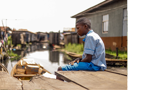 A young boy sitting beside a polluted waterway in an underserved community, representing the health risks of contaminated water and poor sanitation.